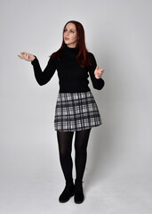 Full length portrait of a red haired  girl wearing black turtleneck sweater,  plaid skirt and boots.  Standing pose against a studio background.