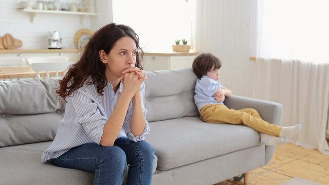 Offended mom and son sit on couch in living room avoid talking and looking at each other after quarrel. Family conflict with young mother or nanny and little disobedient kid at home at covid lockdown