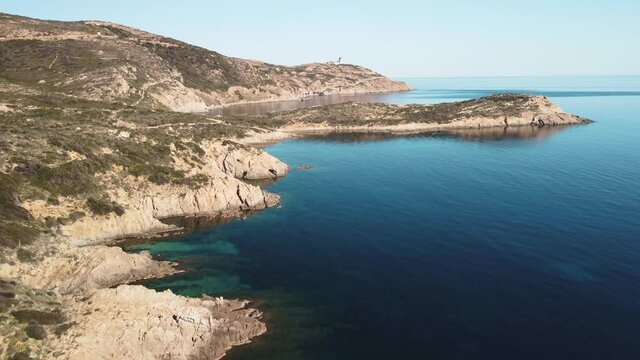 Aerial view of the rocky coastline and turquoise Mediterranean sea of La Revellata near Calvi in the Balagne region of Corsica as a kayak paddles around a shallow bay with a lighthouse in the distance