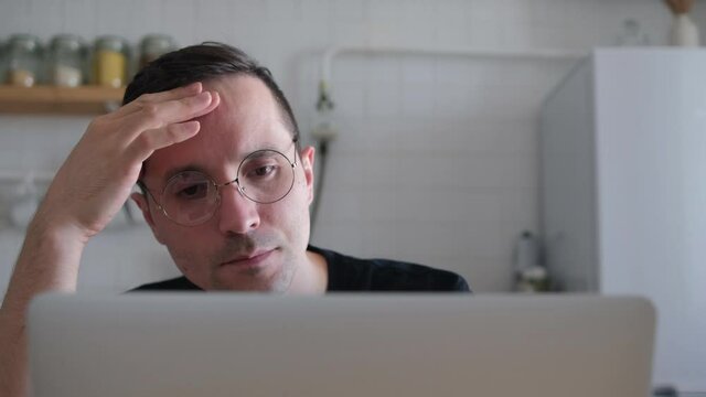 A thoughtful serious young Eupropean student writer sits at a home office desk in the kitchen with a laptop thinking about inspiration finding solutions to problems ideas in thoughts concepts dreams.