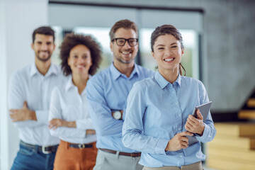 Multicultural group of business people standing with arms crossed in a row in office. Selective focus on woman who is holding tablet.