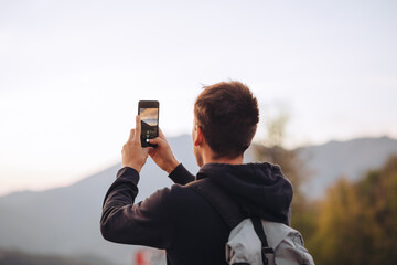 Young guy in a hoodie makes a horizontal photo of sunset on his phone in mountains