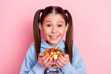 Photo portrait of happy schoolgirl keeping glass with sweet candies isolated on pastel pink color background