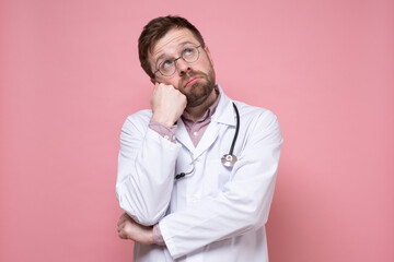 Pensive, sad doctor with a stethoscope around his neck looks up dreamily. Pink background.