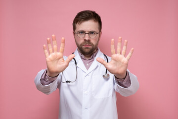 Serious doctor with a stethoscope around neck shows a stop gesture, putting palms forward and looking sternly at the camera. 