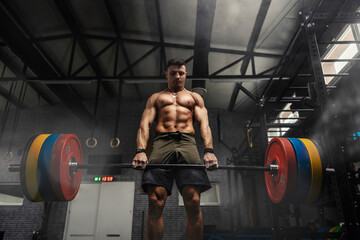 Fototapeta na wymiar Weightlifter at the gym, a moment before a powerful movement. A man with a strong body holds a heavy barbell in his hands and does a dead lift in a dark atmosphere gym. Motivation in sports, cross fit