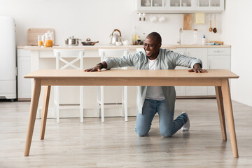 Overjoyed black man touching new wooden table, after assembing furniture with own hands at home in kitchen interior - Powered by Adobe
