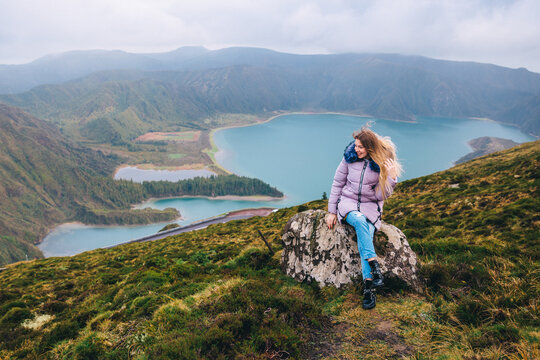 A woman sits on a stone against the backdrop of the beautiful crater of Lake Lagoa do Fogo in the Agua de Pau Massiva stratovolcano in the center of São Miguel Island. Travel to the Azores.