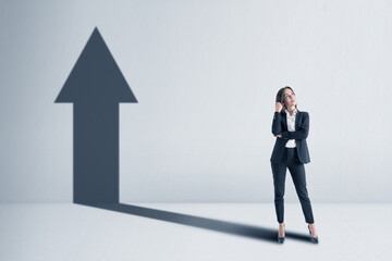 Fototapeta premium Businesswoman with a shadow in the shape of an arrow up on the blank white concrete wall background. Promotion and success concept