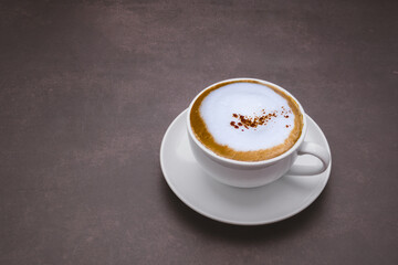 A white coffee cup with a cappuccino on a dark brown background. Space for text. Morning beverage and relaxation concept