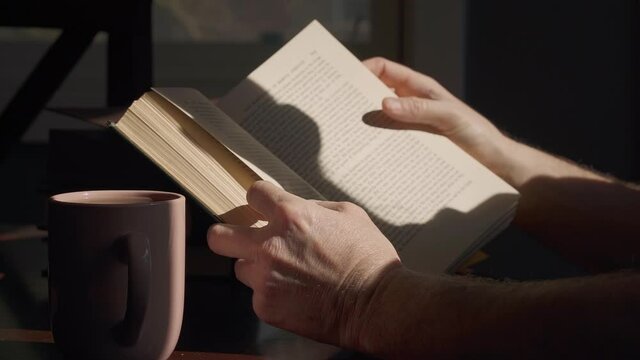 Man near window with natural lighting reading book and drinking coffee