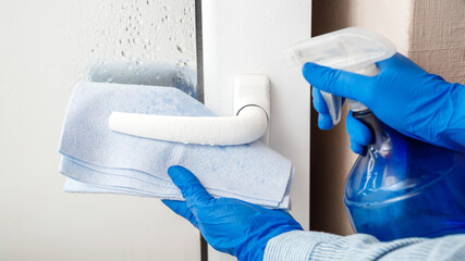 Door handle cleaning by antibacterial alcohol spray. Woman Houseworker in rubber blue gloves clean...