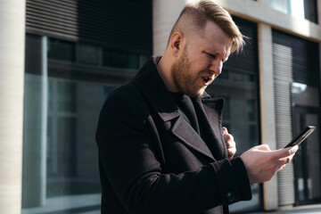 Fototapeta premium A stylish businessman in a black coat holds a phone in his hands and wrinkles his forehead in perplexity