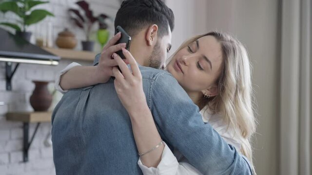 Charming confident young Caucasian woman hugging Middle Eastern man in kitchen messaging online on smartphone. Portrait of absorbed girlfriend surfing social media indoors embracing boyfriend