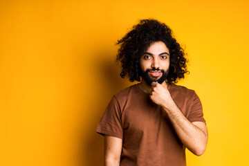 Fototapeta na wymiar Smiling african american guy with a beard and in a brown t-shirt touches his beard while standing on a yellow background