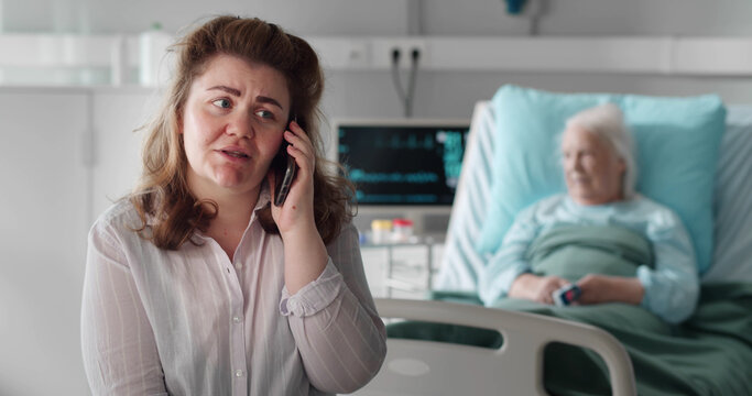 Woman in hospital ward talking on smartphone with sick aged mother in hospital bed on background