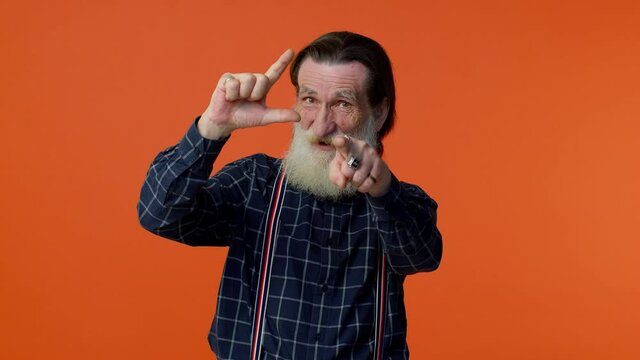 Elderly bearded gray-haired man showing loser gesture and pointing on you, blaming accusing for unsuccess, expressing disrespect, mocking your failure. Senior old grandfather on orange background