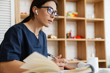 Focused woman doctor using earphone while working with papers at home
