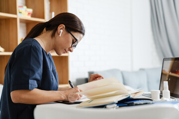 Focused woman doctor using earphone while working with laptop and papers