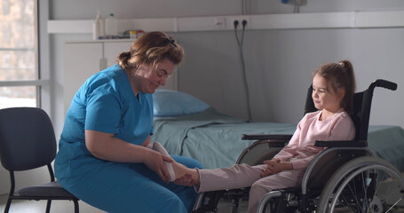 Nurse massaging foot of little girl patient sitting in wheelchair