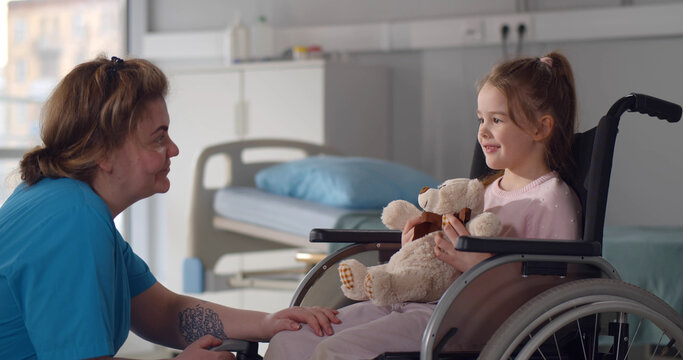 Child patient in wheelchair sitting in hospital ward talking to female nurse