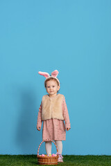 charming smiling little girl stands on a blue background in the studio and holds basket of Easter eggs. Funny happy baby wears rabbit ears on Easter day. Have fun hunting for Easter eggs. Copy space