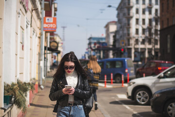 Young cheerful stylish woman using cell phone and texting message on city street