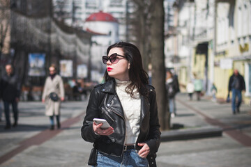 Fototapeta premium Young cheerful stylish woman using cell phone and texting message on city street