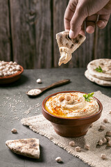 Hand dipping pita bread in hummus on dark background vertical format.