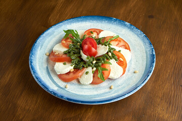 Classic Italian caprese salad with tomatoes, mozzarella, arugula and pesto in a blue plate on a wooden background. Diet salad