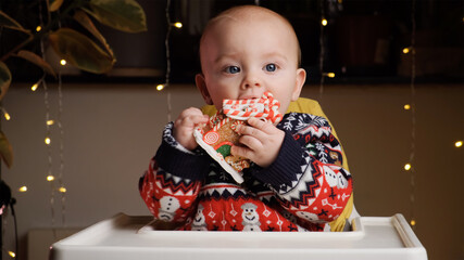 Little baby boy wearing Christmas sweater playing with toy gingerbread house, sitting in baby high chair at home. Christmas night concept.