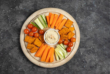 Hummus standing on a wooden plate with sliced cucumbers, carrot sticks, cherry tomatoes and crunchy crackers on a gray background