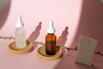 Two glass bottles for cosmetics, natural medicine , essential oils or serum and a piece of handmade soap next to a green tree branch on a white and pink background. Eco friendly beauty products.