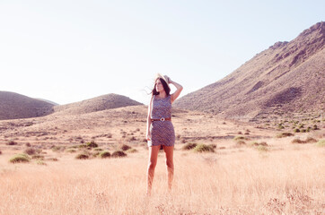 Young woman with long brunette hair holding a hat wears a floral dress in a dry meadow in the late afternoon
