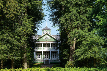 Old green wooden house with a balcony and white columns framed by trees in the manor park.