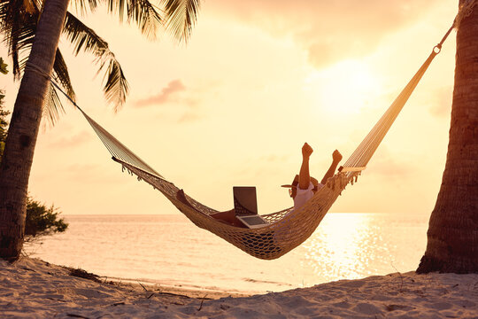 Full length of young woman freelancer celebrating success, keeping arms raised while lying in hammock on the tropical beach at sunset and working on laptop