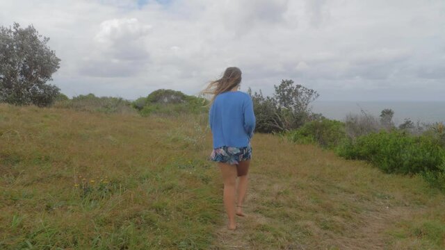 Beautiful Caucasian Woman Walking In Bare Feet On Grassy Hill By The Sea Coast. Big Nobby At Crescent Head Lookout In New South Wales, Australia. rear shot
