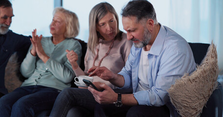 Company of mature men and women gathering inside house to communicate news. Middle-aged old businessman using smartphone checking notifications.