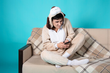 A funny guy is playing on the phone while sitting on the couch. Blue background. Young man in pajamas.