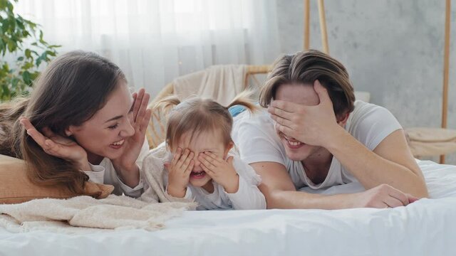 Happy caucasian parents caring mom and loving father playing hide and seek with toddler baby infant child kid daughter girl lying on bed in cozy home, family closes eyes with hands enjoy game together