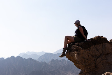 Young female tourist girl sitting at the edge of the rock with abyss below, looking at the mountain...