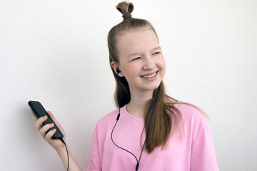 A girl in headphones with a phone in her hand. A young, pretty woman in a simple pink T-shirt looks away and smiles.