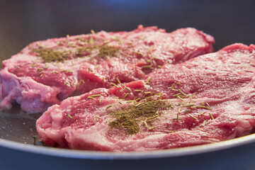 Two red meat steaks with spices in a frying pan.