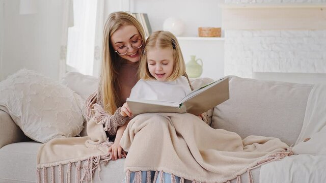 Young mom blonde mother sits with little daughter baby girl on couch covered by warm blanket reading book fairy tale at weekend, mum showing photo album to child kid laughing, talking together at home