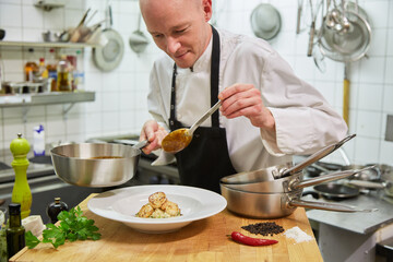 Chef without hair prepares scampi in the kitchen. He holds a pot in his hand and has a spoon in his hand. In the foreground you can see chily, pepper, salt, parsley. Feel like going out for dinner?