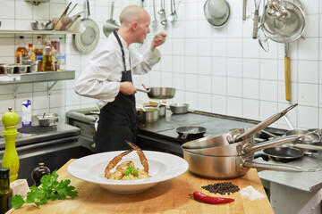 Seafood dish arranged on plate in a kitchen. In the background a cook (without hair, of slender stature) who is seasoning. Foreground: Chilly, parsley, salt, pepper, pans.