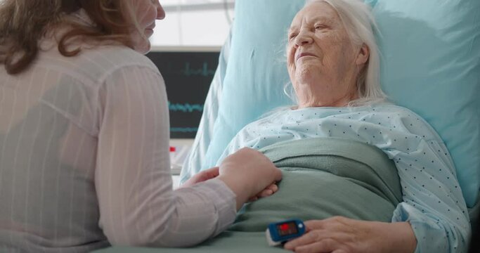 Young daughter visiting and cheering aged mother lying in bed at hospital ward