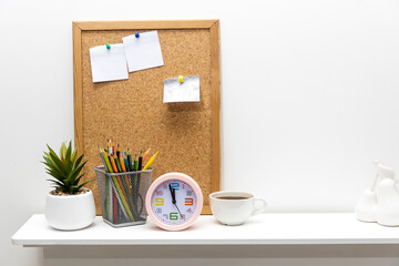 Note board with pinned sheets of paper, artificial succulent in a ceramic pot, clock, pencil holder with pencils on a white table top.