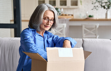 Happy senior middle 60s aged woman opening box with ordered clothes at home on couch. Old mature online shopper customer opening online shop parcel. International delivery service comfort concept. - obrazy, fototapety, plakaty
