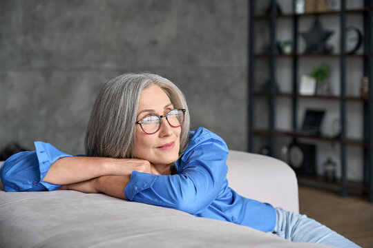 Mature Peaceful 60s Middle Age Lady Sitting Relaxing At Home. Senior Elder Serene Woman In Glasses Looking At Window Thinking Of Positive Vision, Dreaming Of Future And Enjoying Wellbeing.